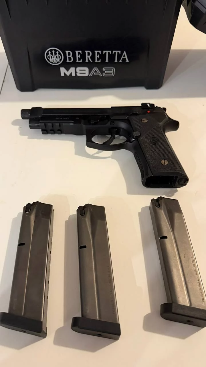 Pistolet Beretta M9A3 noir à canon fileté posé sur une table blanche, trois chargeurs devant et sa caisse derrière.