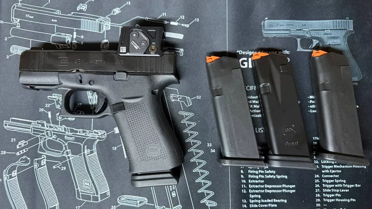 Pistolet Glock 43X noir avec point rouge sur la culasse, posé sur un tapis d'entretien à côté de trois chargeurs à élévateur orange.