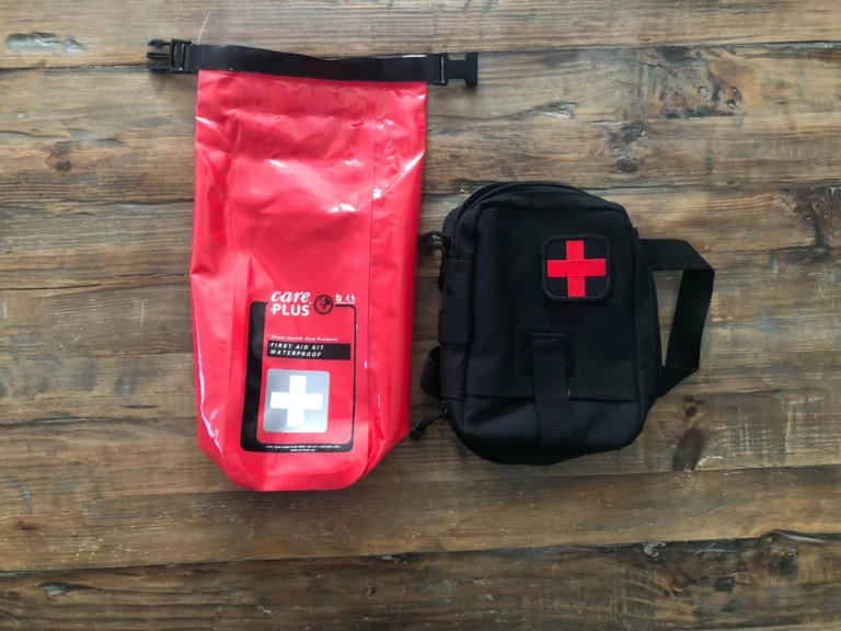 Kit de secours Care Plus dans son sac étanche rouge, à côté d'une pochette noire marquée d'une croix rouge