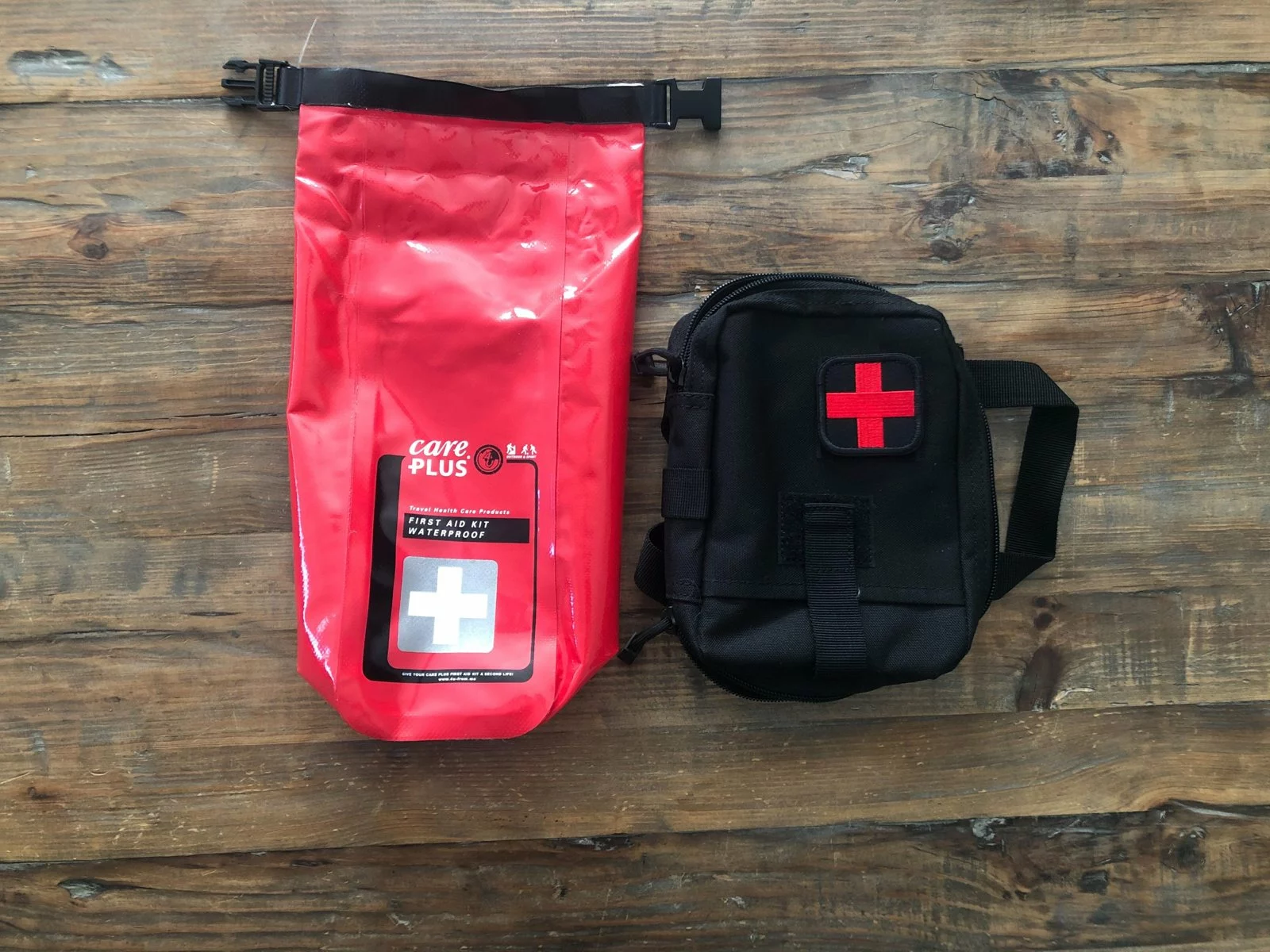 Kit de secours Care Plus dans son sac étanche rouge, à côté d'une pochette noire marquée d'une croix rouge